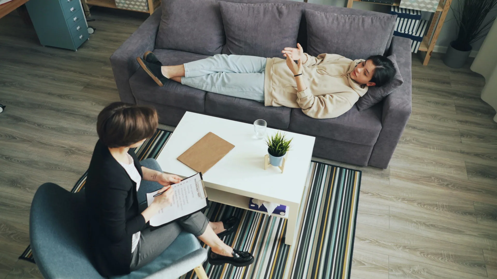 A young individual is quietly talking about their feelings while relaxing on a sofa. A counsellor is taking notes on the situation.